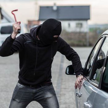 Man dressed in black with a balaclava on his head breaking a glass in car with crowbar. Car thief car theft concept
